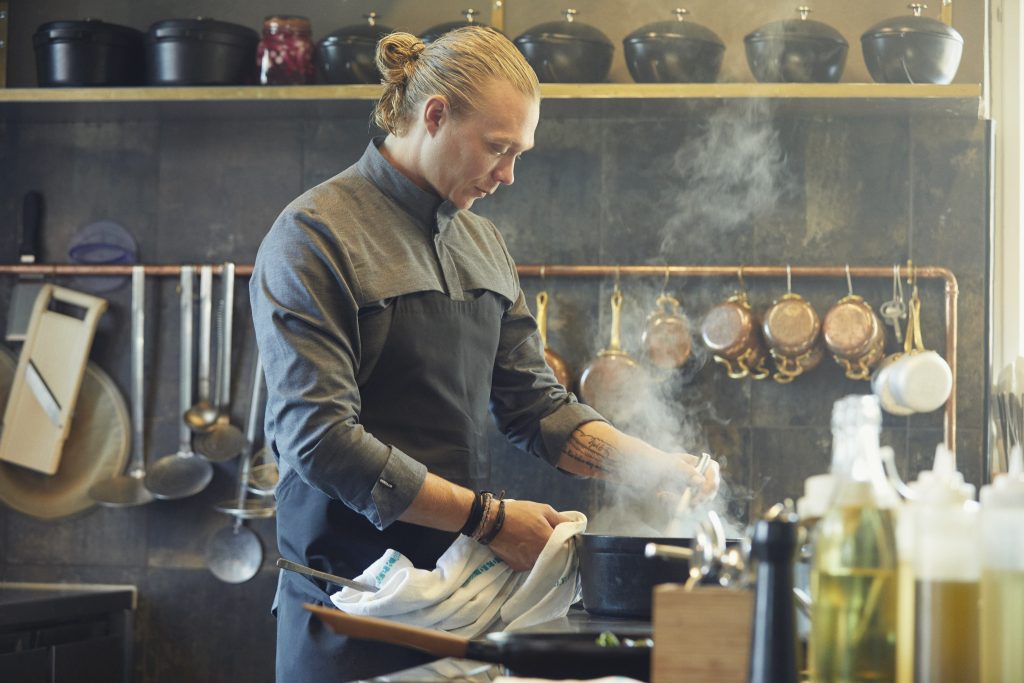 Ein junger Koch trägt die funktionelle und gut durchdachte Workwear von Kentaur. Kochhemd in dunkelgrau und schwarze Schürze. Aus dem Topf vor ihm steigt Dampf auf.